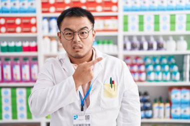 Chinese young man working at pharmacy drugstore pointing aside worried and nervous with forefinger, concerned and surprised expression 