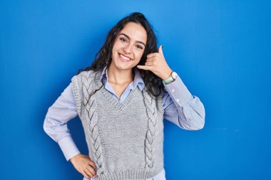 Mavi arka planda durup gülümseyen esmer bir kadın telefonla el ve parmaklarıyla telefonla konuşur gibi hareket ediyor. iletişim kavramları. 