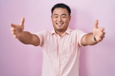 Chinese young man standing over pink background looking at the camera smiling with open arms for hug. cheerful expression embracing happiness. 