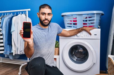 Middle east man with beard showing smartphone screen and washing machine clueless and confused expression. doubt concept. 