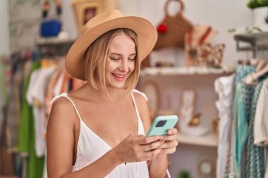 Young blonde woman customer wearing summer hat using smartphone at clothing store