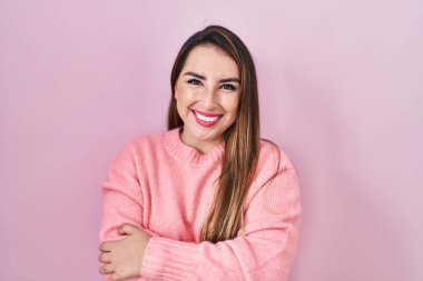 Young hispanic woman standing over pink background happy face smiling with crossed arms looking at the camera. positive person. 