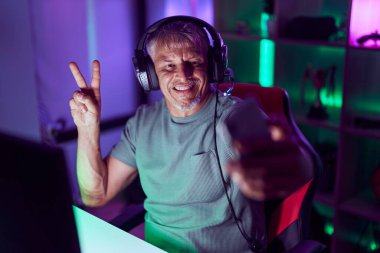 Orta yaşlı, kır saçlı erkek flamalı. Oyun odasında akıllı telefondan selfie çekiyor.