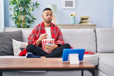 Young hispanic man eating popcorn watching movie on tablet device looking at the camera blowing a kiss being lovely and sexy. love expression. 