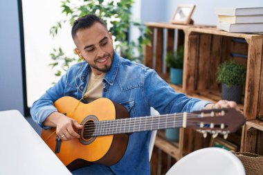 Young hispanic man playing classical guitar sitting on table at home