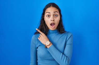 Mavi izole edilmiş arka planda dikilen genç Brezilyalı kadın parmağını yana çevirerek şaşırdı, ağzı açık hayret içinde kaldı.. 