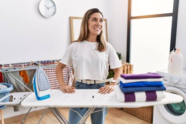 Young hispanic woman ironing clothes at laundry room looking away to side with smile on face, natural expression. laughing confident. 