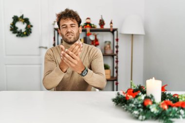 Masada sakallı genç yakışıklı bir adam Noel süslemesi ile oturuyor. Ellerinde ve parmaklarında ağrı var. Eklem iltihabı var. 
