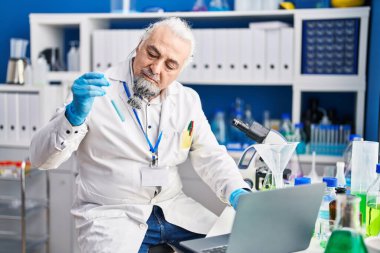 Middle age grey-haired man scientist using laptop holding test tube at laboratory