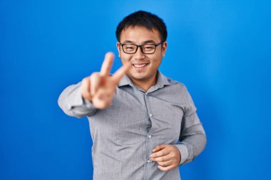 Young chinese man standing over blue background smiling looking to the camera showing fingers doing victory sign. number two. 