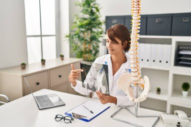 Young beautiful hispanic woman doctor pointing with finger to vertebral column xray at clinic