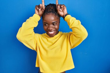 Beautiful black woman standing over blue background doing funny gesture with finger over head as bull horns 