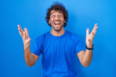 Hispanic young man standing over blue background celebrating crazy and amazed for success with arms raised and open eyes screaming excited. winner concept 