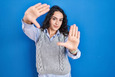 Genç esmer kadın mavi arka planda duruyor. El ve parmakları kullanarak çerçeve yapıyor. Kamera bakış açısı. 