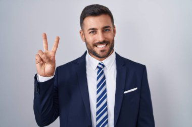 Yakışıklı İspanyol takım elbiseli ve kravatlı adam iki numaralı parmaklarını gösterip gösterirken kendinden emin ve mutlu gülümsüyor.. 