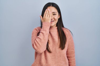 Young latin woman standing over blue background covering one eye with hand, confident smile on face and surprise emotion. 