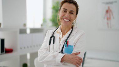 Orta yaşlı İspanyol kadın doktor üniforması giyiyor klinikte kollarını kavuşturup el kol hareketi yapıyor.