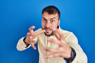 Handsome hispanic man standing over blue background shouting frustrated with rage, hands trying to strangle, yelling mad 