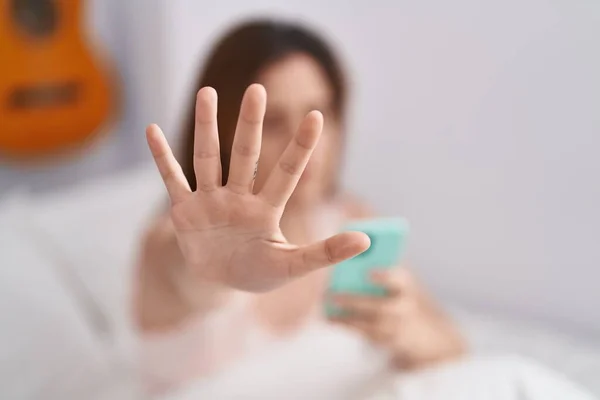Young woman using smartphone doing stop gesture with hand at bedroom