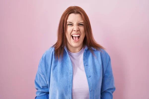 Young hispanic woman with red hair standing over pink background angry and mad screaming frustrated and furious, shouting with anger. rage and aggressive concept.