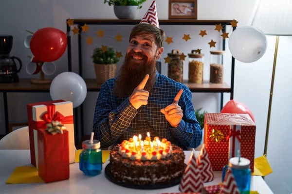 白种人蓄着长胡子庆祝生日 微笑着拿着大巧克力蛋糕 看着摄像机 用双手和手指指向旁边 — 图库照片