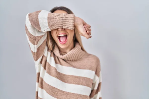Young blonde woman wearing turtleneck sweater over isolated background covering eyes with arm smiling cheerful and funny. blind concept.