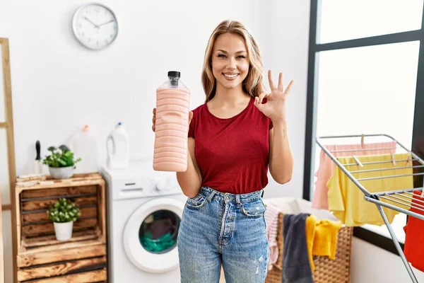 年轻的高加索女人在洗衣房拿着洗涤剂瓶子 用手指做了个手势 微笑着友好地摆设着极好的标志 — 图库照片