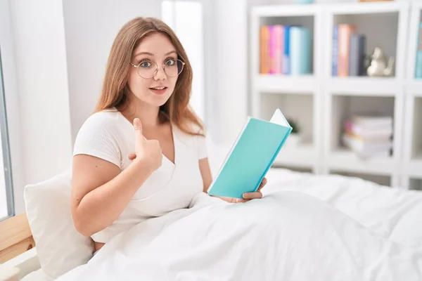 Young caucasian woman sitting on the bed at home reading a book pointing finger to one self smiling happy and proud
