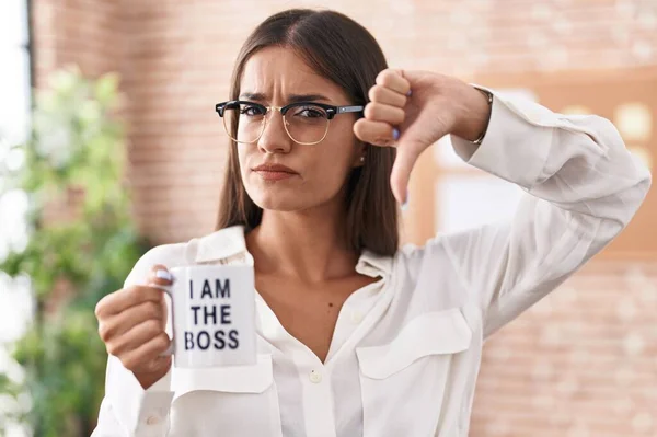 Young brunette woman drinking from i am the boss coffee cup with angry face, negative sign showing dislike with thumbs down, rejection concept
