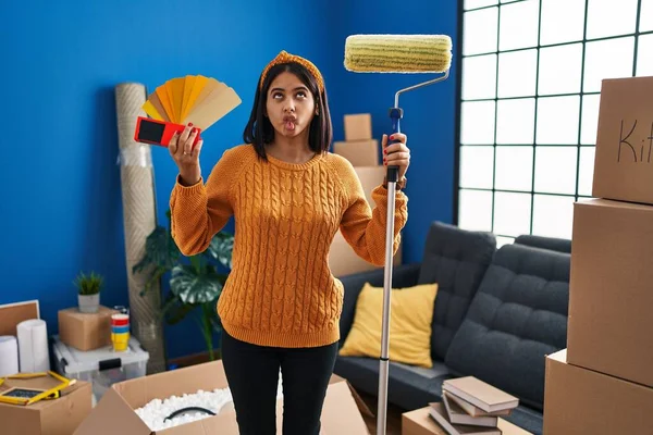 Young hispanic woman painting home walls with paint roller making fish face with mouth and squinting eyes, crazy and comical.