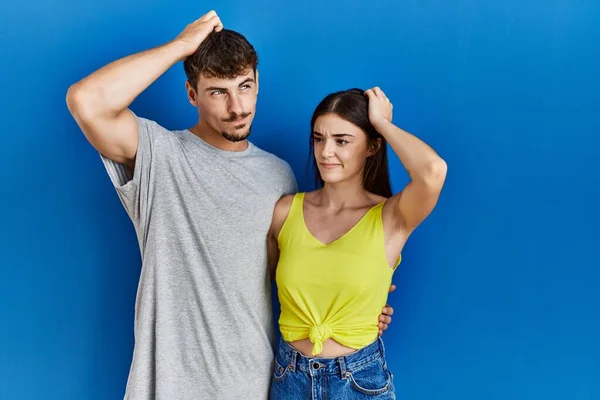 Young hispanic couple standing together over blue background confuse and wondering about question. uncertain with doubt, thinking with hand on head. pensive concept.