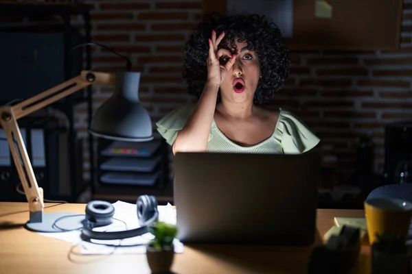 Young brunette woman with curly hair working at the office at night doing ok gesture shocked with surprised face, eye looking through fingers. unbelieving expression.
