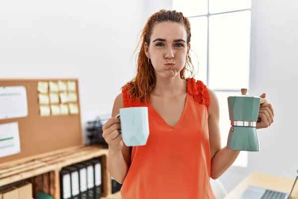 Young redhead woman holding french coffee maker at the office puffing cheeks with funny face. mouth inflated with air, catching air.