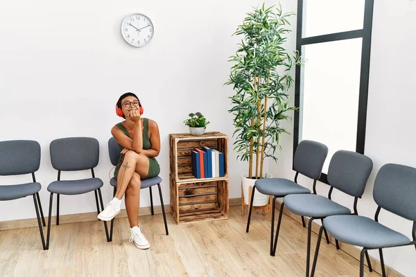 Young hispanic woman listening to music sitting on chair at waiting room