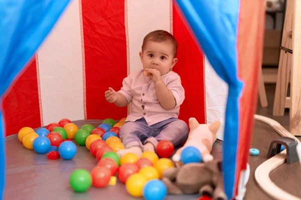 Adorable toddler bitting plastic construction blocks sitting inside of circus tent at home