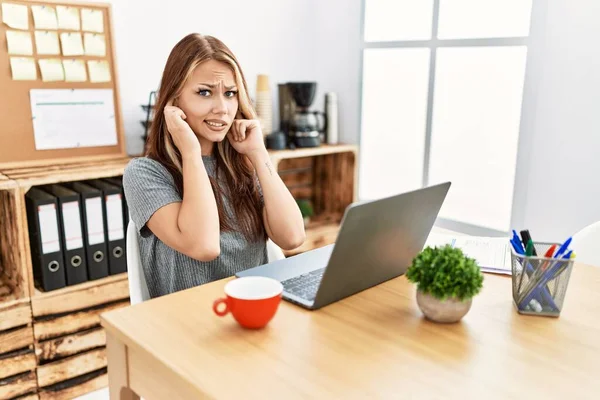 Young brunette woman working at the office with laptop covering ears with fingers with annoyed expression for the noise of loud music. deaf concept.