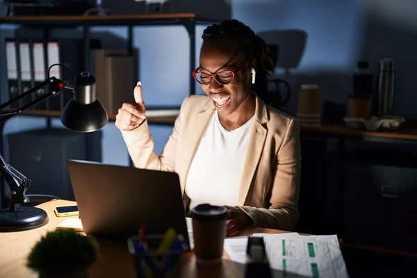 Beautiful black woman working at the office at night doing happy thumbs up gesture with hand. approving expression looking at the camera showing success.