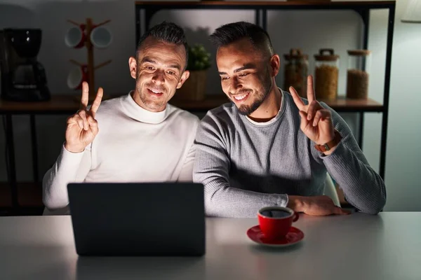 Homosexual couple using computer laptop smiling with happy face winking at the camera doing victory sign with fingers. number two.