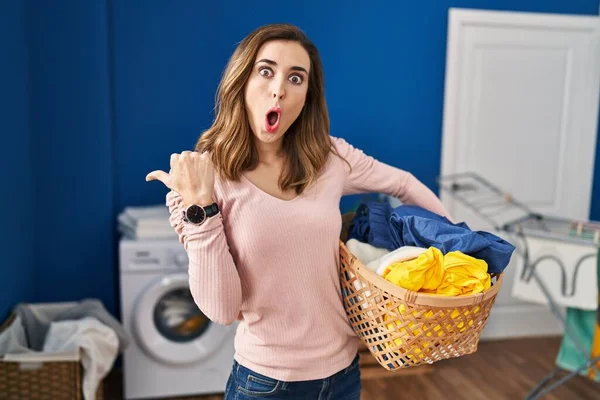 Young woman holding laundry basket surprised pointing with hand finger to the side, open mouth amazed expression.