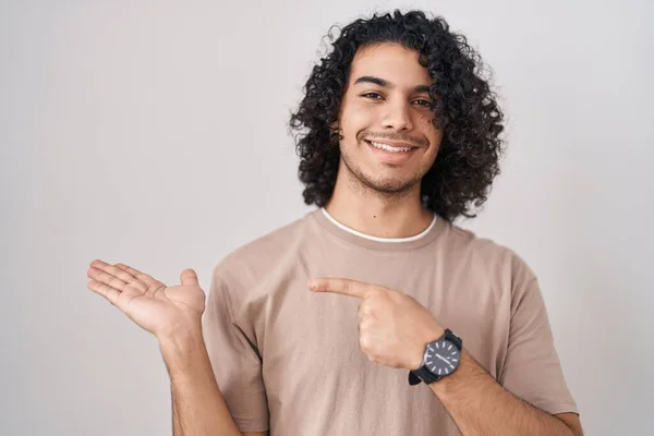 头发卷曲的西班牙裔男子站在白色的背景上 一边手拿着相机 一边用手指指指着 一边惊讶地微笑着 — 图库照片