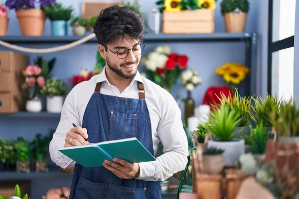 Young hispanic man florist smiling confident writing on notebook at florist shop