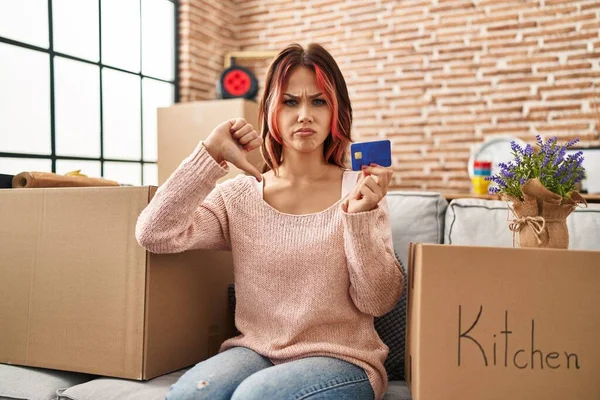 若い白人女性が怒っている顔をしたクレジットカードを持って新しい家に移動します 親指で嫌いを示す負の記号ダウン 拒絶概念 — ストック写真