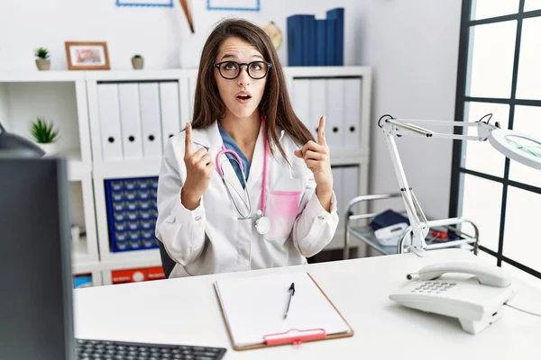 Young doctor woman wearing doctor uniform and stethoscope at the clinic amazed and surprised looking up and pointing with fingers and raised arms.