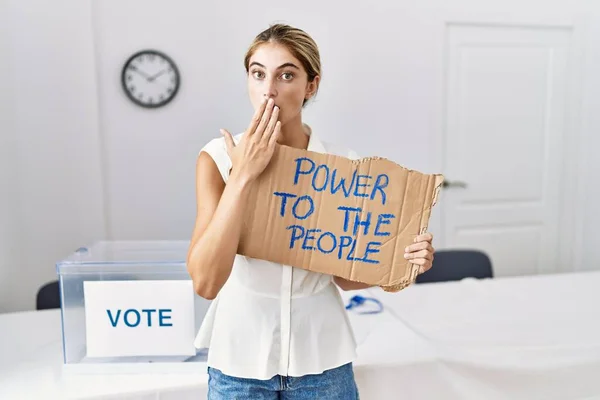 Young blonde woman at political election holding power to the people banner covering mouth with hand, shocked and afraid for mistake. surprised expression