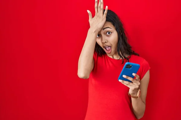 Young brazilian woman using smartphone over red background surprised with hand on head for mistake, remember error. forgot, bad memory concept.