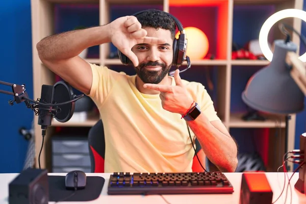 Hispanic man with beard playing video games with headphones smiling making frame with hands and fingers with happy face. creativity and photography concept.