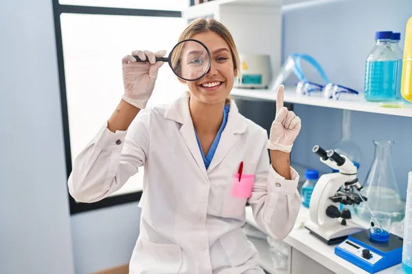Young blonde woman working at scientist laboratory using magnifying glass smiling with an idea or question pointing finger with happy face, number one