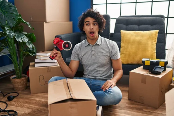 Hispanic man with curly hair moving to a new home closing cardboard box with tape scared and amazed with open mouth for surprise, disbelief face