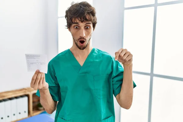 Young hispanic doctor man holding covid record card pointing down with fingers showing advertisement, surprised face and open mouth