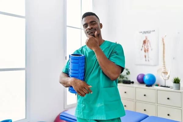 Young African Physiotherapist Man Holding Foam Roller Clinic Serious Face — Foto de Stock